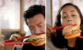 Hình ảnh nhân vật ăn bánh burger bằng đôi đũa màu đỏ cỡ lớn. (Ảnh chụp từ clip)