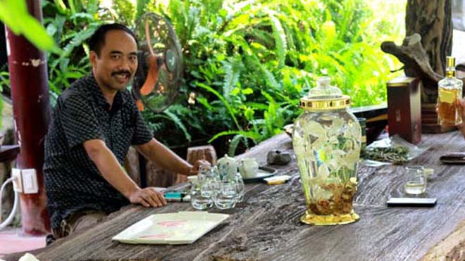 Anh Tuyền ngồi ngắm bình rượu sâm trên chiếc bàn gỗ hóa thạch khổng lồ, giữa khu vườn đầy hoa lá, chim muông 