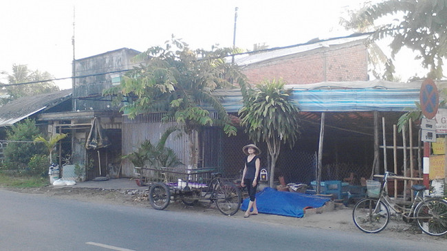 Bà Nguyệt đứng trước dãy nhà xập xệ vì đất tranh chấp hơn 30 năm, bên con đường vừa tráng nhựa. Ảnh: Sáu Nghệ.