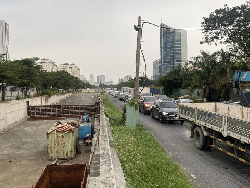 Phía bên trái là công trường, phía phải là cảnh xe tải, ô tô, container xếp hàng dài di chuyển qua khu vực này.