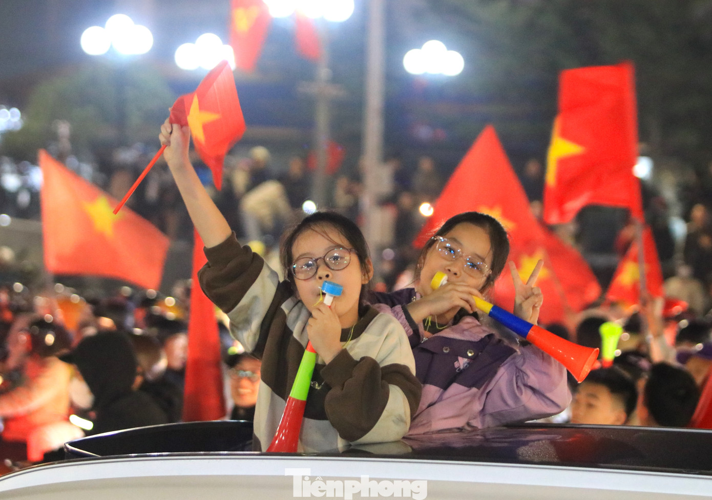 2 bé gái xinh xắn đứng trên cửa sổ trời ô tô phất cờ, thổi còi cùng ăn mừng chức vô địch của đội tuyển Việt Nam.