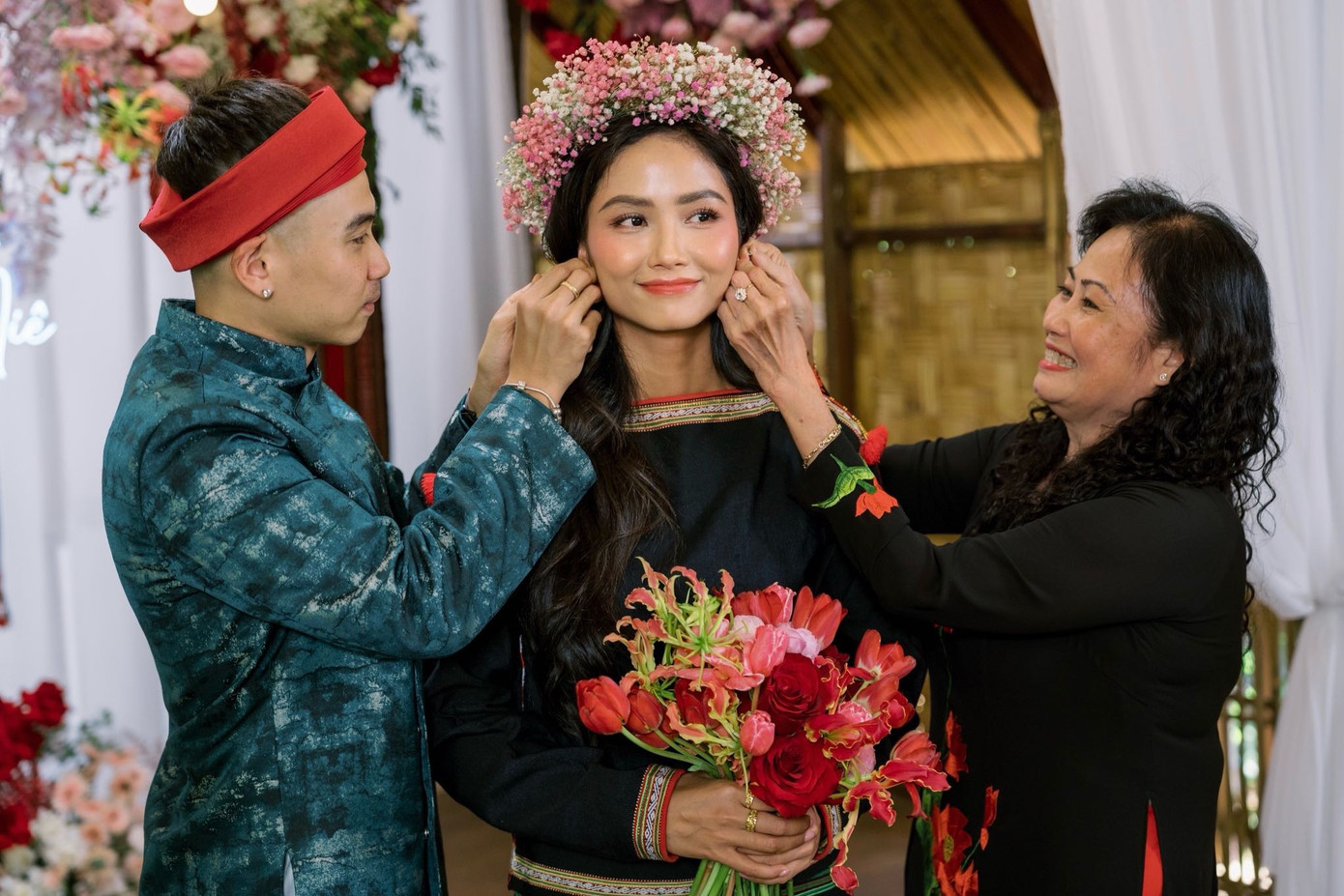 Tuấn Khôi và mẹ chồng trao trang sức cho H&apos;Hen Niê. Cô dâu đi chân đất khi diện trang phục truyền thống của người Ê-Đê và đội vòng hoa baby với tông trắng, hồng chủ đạo.