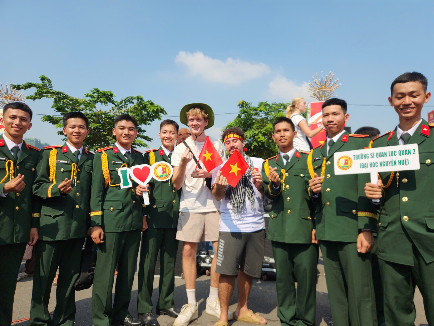 Du khách Tây đội nón tai bèo, cầm cờ đỏ sao vàng, quấn khăn rằn và băng đô khẩu hiệu Tôi yêu Việt Nam chụp ảnh cùng sĩ quan Lục quân.