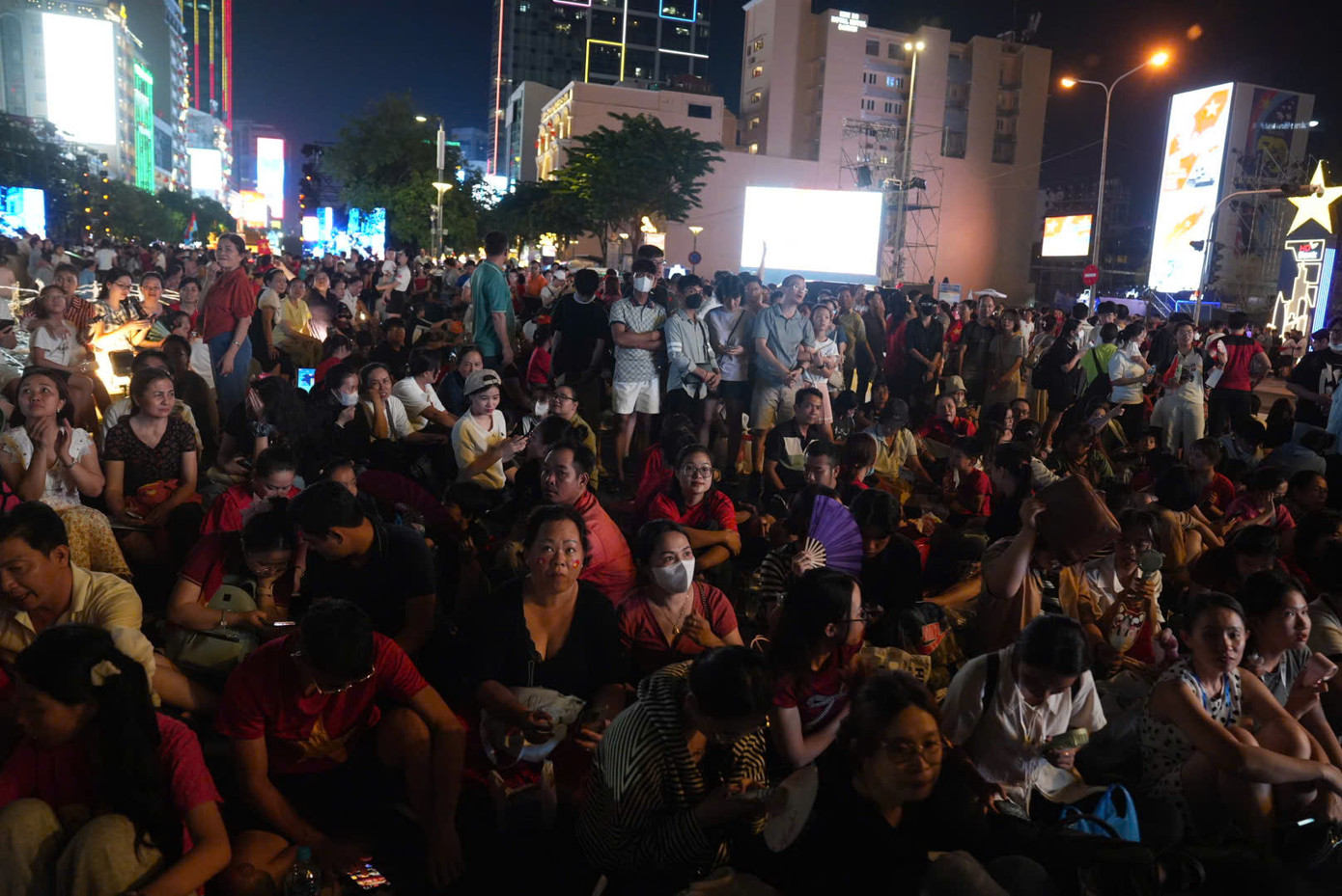 Từng nhóm gia đình ngồi chen sát nhau trước tòa nhà. Nhiều người mang theo quạt tay, nước uống, giữ vị trí từ sớm để thưởng thức trọn vẹn chương trình.