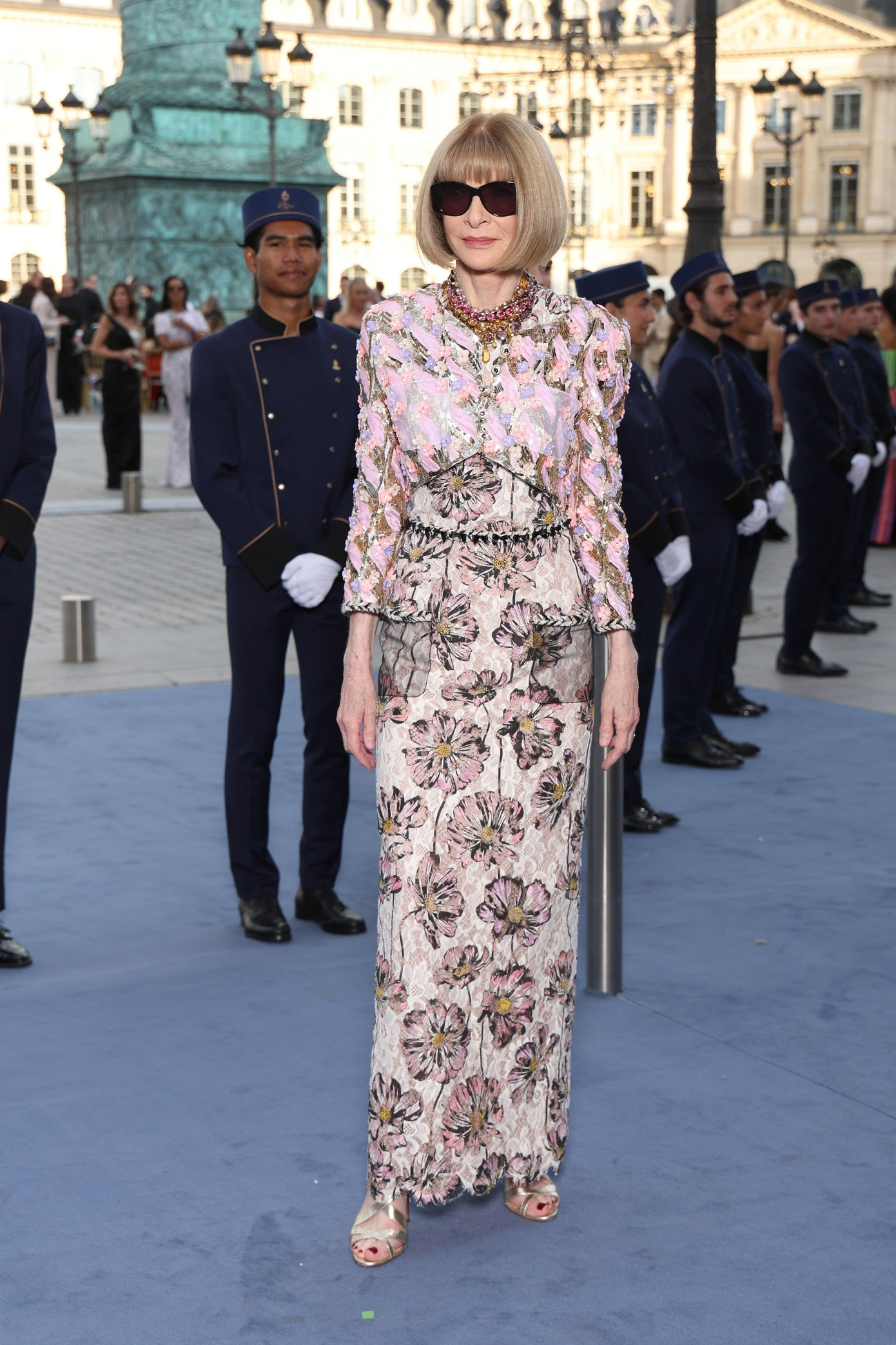 Anna Wintour xuất hiện với mái tóc bob quen thuộc. Tổng biên tập Vogue Mỹ chơi trội khi phối trang phục họa tiết hoa với áo khoác chất liệu phản quang. Nhà báo 74 tuổi là một trong những nhân vật mặc phá cách nhất tại sự kiện.