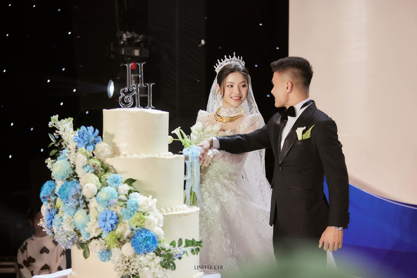 Nghi thức cắt bánh không thể thiếu trong tiệc cưới của những cặp đôi mới cưới. Bánh cưới ba tầng của Quang Hải - Chu Thanh Huyền cao gần 1 m, trang trí cầu kỳ bằng hoa tươi. Đỉnh bánh đính chữ H&amp;H là tên viết tắt của hai người. Trong ảnh, cô dâu và chú rể nhìn nhau trìu mến trong giây phút cùng cắt bánh.