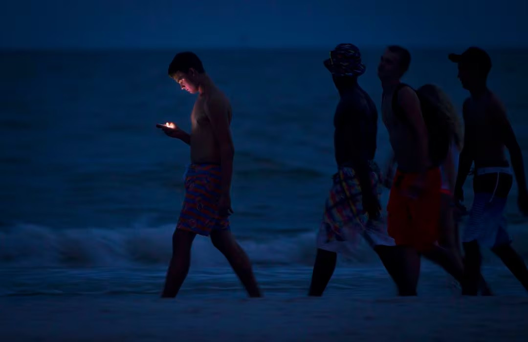 Một người trẻ nhìn vào màn hình điện thoại di động của mình khi đi dạo dọc bãi biển Ft Myers ở bang Florida, Mỹ lúc chạng vạng. Ảnh: Reuters.