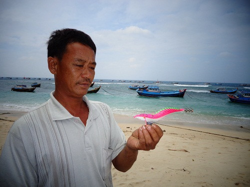 6. Ngư dân Nguyễn Văn Như, 41 tuổi, chủ tàu câu mực đang cầm trên tay con tôm giả dùng làm mồi câu mực. Theo nghề câu mực từ năm 12 tuổi, đến nay anh Như đã là một ngư phủ lão luyện. Mỗi chuyến thả câu cách bờ khoảng 17 hải lý, từ 2 giờ sáng tới 4 giờ chiều, anh câu được 20 – 25kg mực, bán được 3 triệu đồng, trừ chi phí xăng dầu…còn khoảng 2,5 triệu. Tuy vậy, để nuôi vợ và 3 con gái, trong đó con gái đầu đang học Trường cao đẳng mầm non ở Nha Trang, anh Như cho biết chỉ dành dụm được vài chục triệu vì một năm chỉ đi biển được 6 tháng. Như hôm nay biển động, tàu anh phải nằm bờ