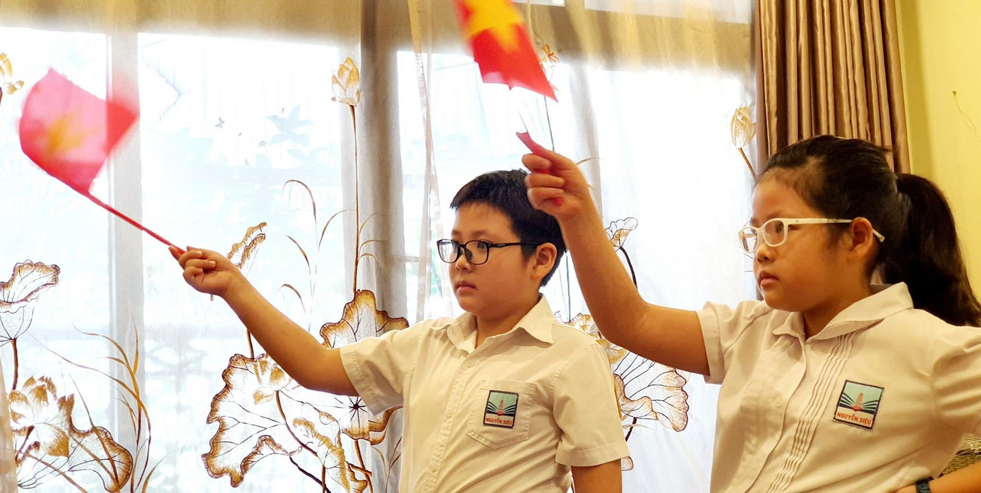 Trần Khởi Nguyên lớp 5ci6 và Trần Quế Linh lớp 3ci4 trường tiểu học Nguyễn Siêu tham dự lễ khai giảng trực tuyến chào cờ trước màn hình tivi tại nhà