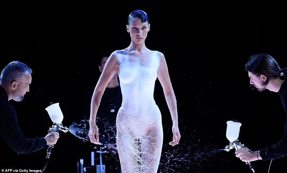 Hai người đàn ông có nhiệm vụ phun lớp mủ màu trắng lên người Bella tạo ra kết cấu thay cho vải ôm lấy đường cong cơ thể. Trong lúc này, Bella đứng yên và điều chỉnh cánh tay vừa tạo dáng biểu diễn vừa thuận tiện cho thành viên ê-kíp làm việc. Ảnh: Getty Images.