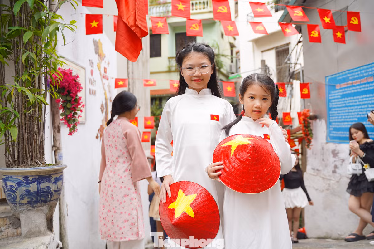 Hai em bé mặc áo dài cầm trên tay nón lá hình Quốc kỳ chụp hình tại ngõ cờ hoa Yên Phụ.