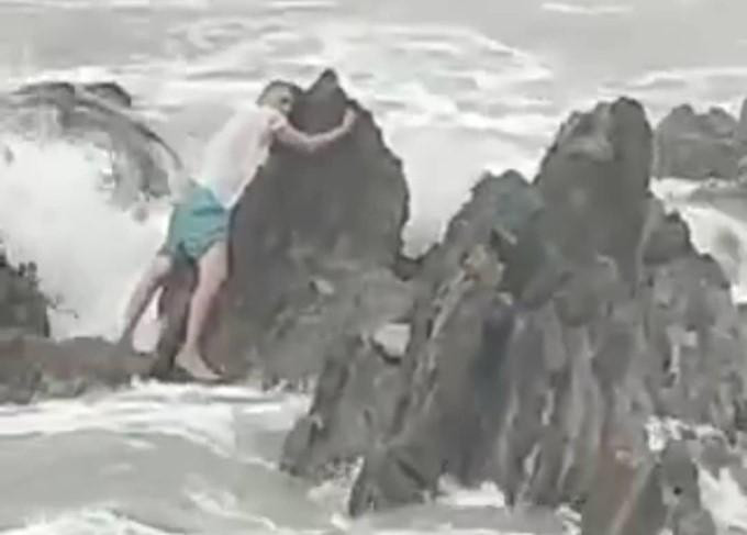 Người đàn ông mải mê chụp ảnh bị sóng biển cuốn ra xa nhưng đã bám được vào tảng đá nhô cao. Ảnh cắt từ clip.