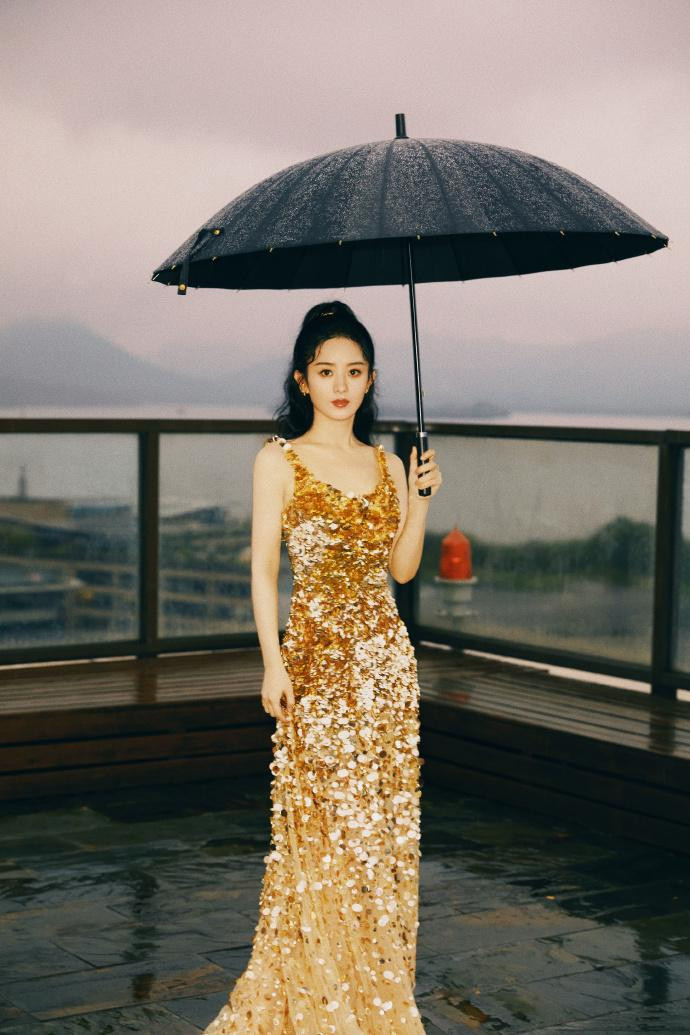 Triệu Lệ Dĩnh mặc váy dạ hội đính sequin vàng lấp lánh. Mái tóc đen gợn sóng buộc kiểu đuôi ngựa, đeo bông tai "ton-sur-ton" với trang phục.