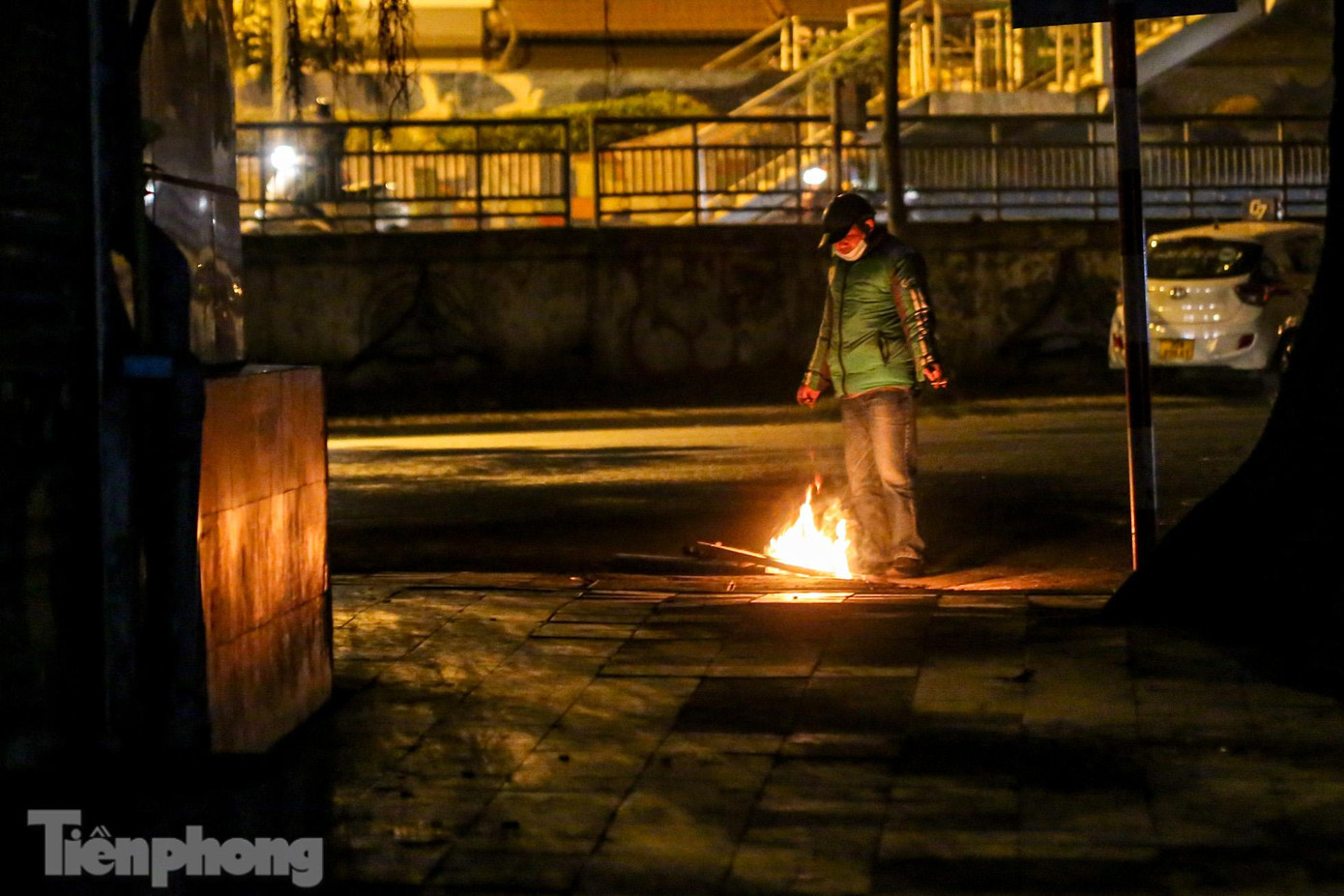 Một tài xế xe ôm công nghệ đang đứng cạnh đống lửa, vừa chờ khách, vừa thêm củi vào để lửa không bị tắt.