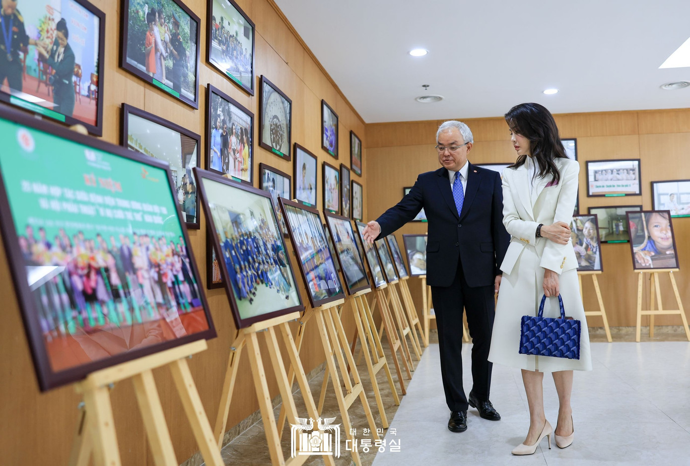 Phu nhân Tổng thống Hàn Quốc phối áo vest dài tay cúc vàng màu trắng với chân váy đồng bộ và áo sơ mi trắng cổ cồn, kết hợp giày cao gót màu be và túi xách da táo thân thiện với môi trường. Set đồ cùng kiểu tóc buông xõa và kiểu trang điểm nhẹ nhàng tạo nên diện mạo trang nhã, sang trọng mà vẫn thoải mái, phù hợp mọi hoàn cảnh.