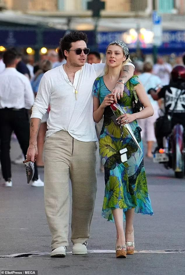 Người mẫu 27 tuổi mặc váy xanh in hoa mỏng tang lộ nội y đen sexy. Cô hoàn thiện vẻ ngoài nữ tính với khăn đội đầu, túi kẹp nách và đôi sandal đế &quot;bánh mì&quot;. Trong khi đó, cậu cả nhà Beckham mặc áo vải lanh trắng kết hợp với quần tây ống rộng màu be và giày thể thao đồng bộ. Phụ kiện đi kèm là mắt kính đen, vòng cổ, đồng hồ và nhẫn cưới.