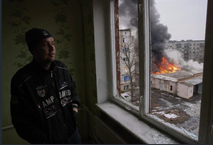 Một người đàn ông nhìn ra cửa sổ, nơi đang có đám cháy sau vụ pháo kích ở Donetsk ngày 27/2. (Ảnh: AP)