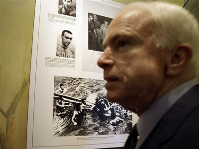 Ông John McCain bên những bức ảnh trong khu di tích nhà tù Hỏa Lò, bức hình bên dưới chụp cảnh quân, dân Hà Nội đang dìu ông từ hồ Trúc Bạch lên bờ vào ngày 26 tháng 10 năm 1967 sau khi máy bay của ông bị bắn hạ. Ảnh AP.