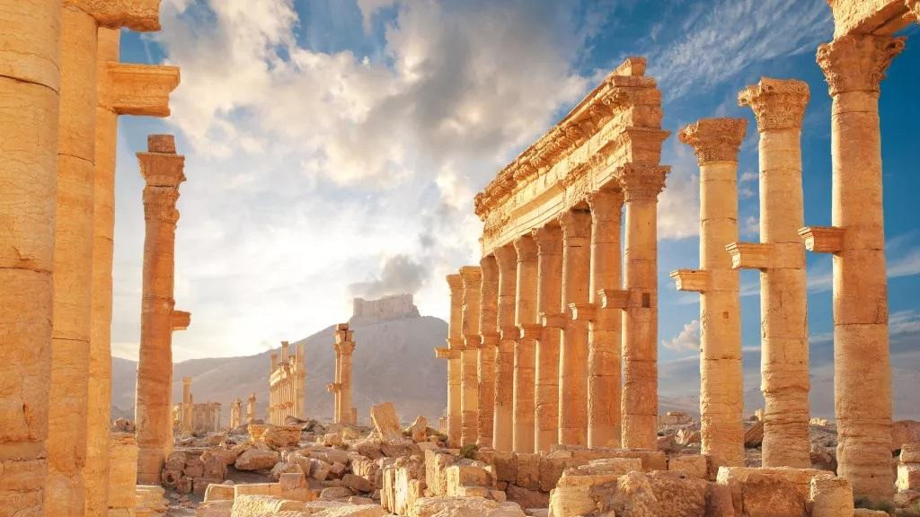 Thị trấn thời La Mã cổ đại ở Palmyra, Syria. Phần lớn bị phá hủy, chỉ còn một số cột đứng sừng sững, ở phía sau là một ngọn đồi với một khu định cư trên đỉnh trên nền trời xanh, có mây.Thành phố cổ Palmyra phát triển rực rỡ cách đây khoảng 2.000 năm như một trung tâm thương mại. Thị trấn thời La Mã cổ đại ở Palmyra, Syria. Phần lớn bị phá hủy, chỉ còn một số cột đứng sừng sững, ở phía sau là một ngọn đồi với một khu định cư trên đỉnh trên nền trời xanh, có mây.Thành phố cổ Palmyra phát triển rực rỡ cách đây khoảng 2.000 năm như một trung tâm thương mại.