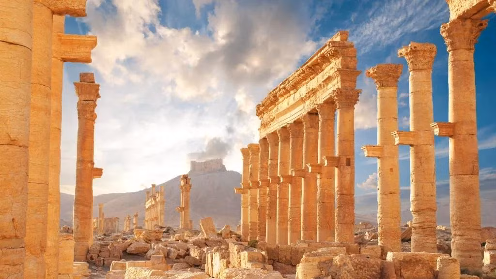 Thị trấn thời La Mã cổ đại ở Palmyra, Syria. Phần lớn bị phá hủy, chỉ còn một số cột đứng sừng sững, ở phía sau là một ngọn đồi với một khu định cư trên đỉnh trên nền trời xanh, có mây.Thành phố cổ Palmyra phát triển rực rỡ cách đây khoảng 2.000 năm như một trung tâm thương mại.