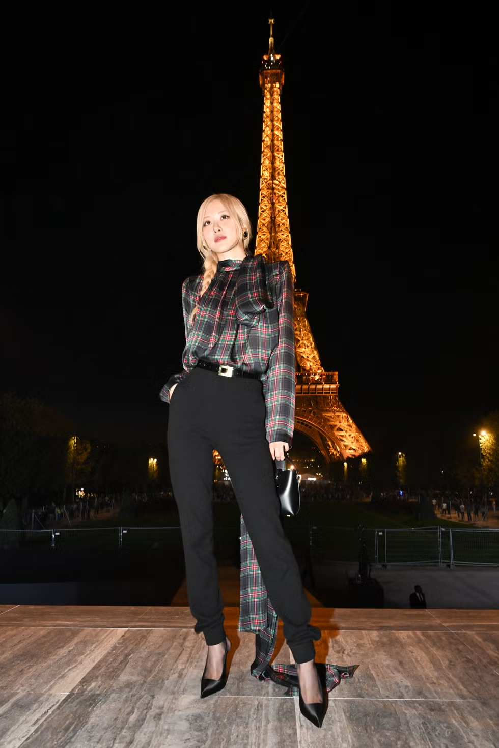 Tạo dáng với Tháp Eiffel trong chiếc áo cà vạt kẻ caro và quần dài đen trong chuyến tham dự buổi trình diễn Xuân/Hè 2024 của Saint Laurent.