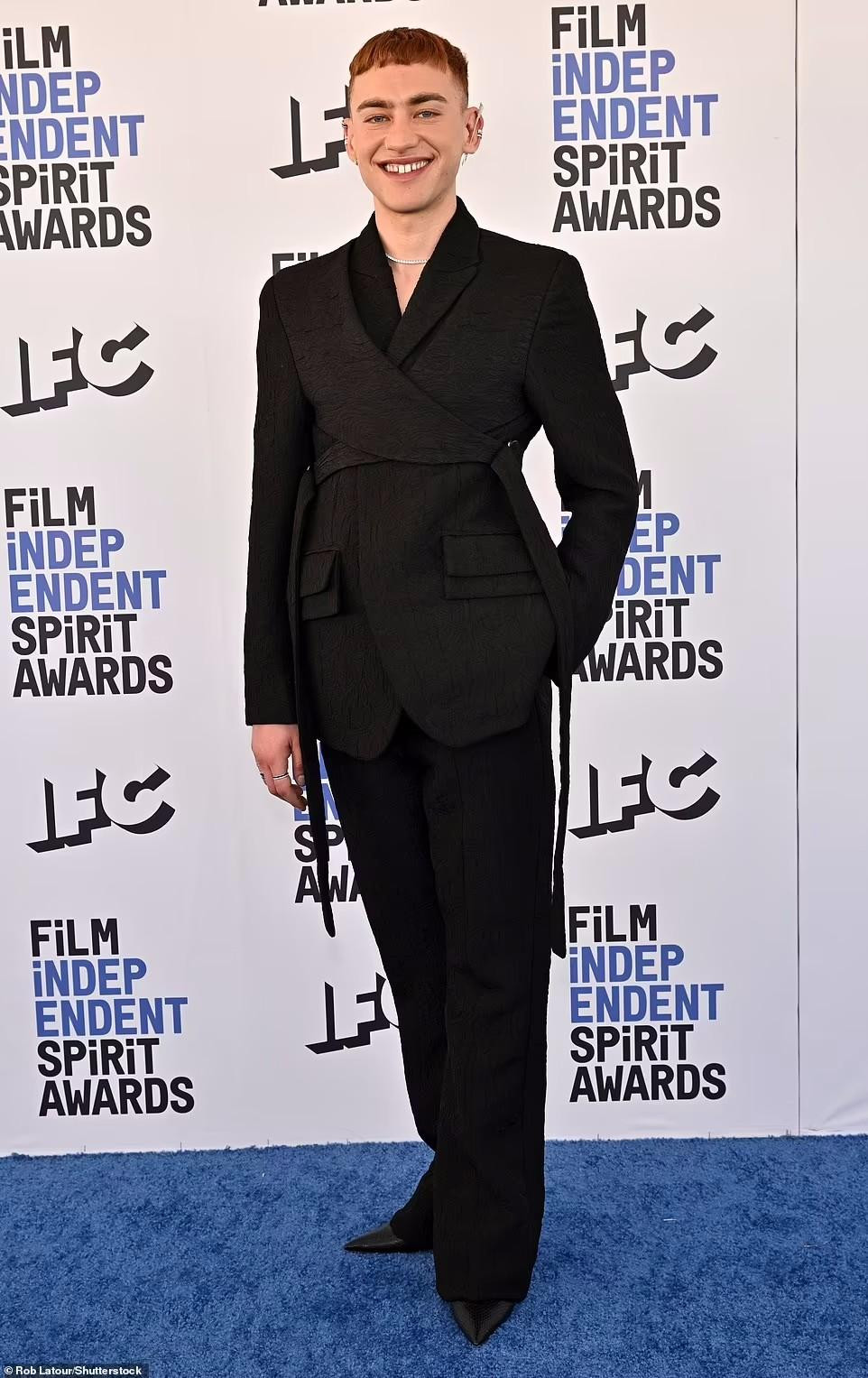 Ca sĩ người Anh Olly Alexander mặc một bộ quần áo màu đen với bông tai mũi tên bạc.