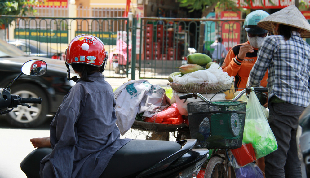 Một em bé được mặc áo của mẹ, ngồi chờ mẹ mua hoa quả. Ảnh: Trường Phong