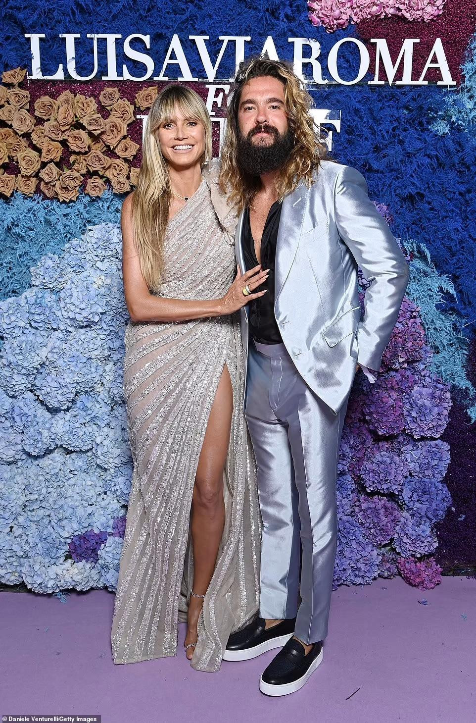 Heidi Klum diện đầm bạc lệch vai lấp lánh sánh đôi cùng ông xã kém 16 tuổi Tom Kaulitz - nổi bật với bộ vest bóng lộn. Thiết kế xẻ tà cao giúp siêu mẫu Mỹ gốc Đức khoe chân thon dài gợi cảm. Khó có thể tin người đẹp đã 48 tuổi. Cô thậm chí còn trẻ hơn chồng.