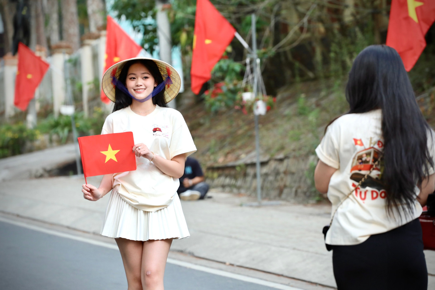Bạn trẻ khoác lên mình chiếc áo với dòng chữ Độc Lập - Tự Do - Hạnh Phúc và đội nón lá truyền thống check-in con đường cờ hoa.