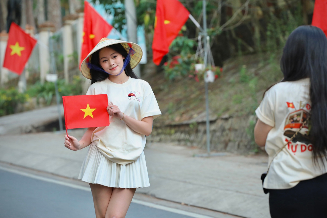 Bạn trẻ khoác lên mình chiếc áo với dòng chữ Độc Lập - Tự Do - Hạnh Phúc và đội nón lá truyền thống check-in con đường cờ hoa.