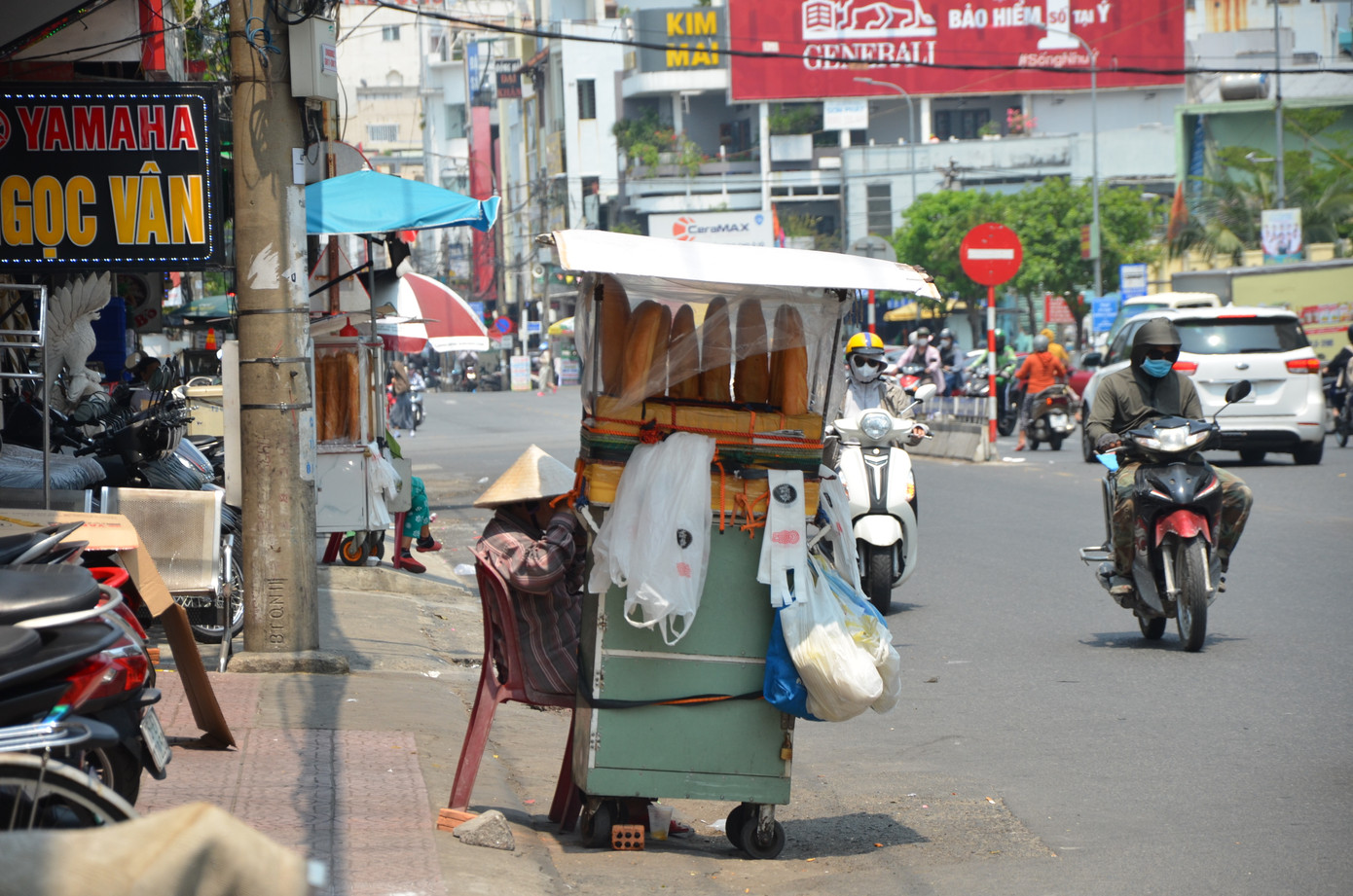 Tại đường Hùng Vương, những người bán bánh mì dạo vốn đã khó khăn nay càng vất vả hơn. Giữa trời nắng gần 40 độ C, họ chỉ có tấm bìa để che nắng.