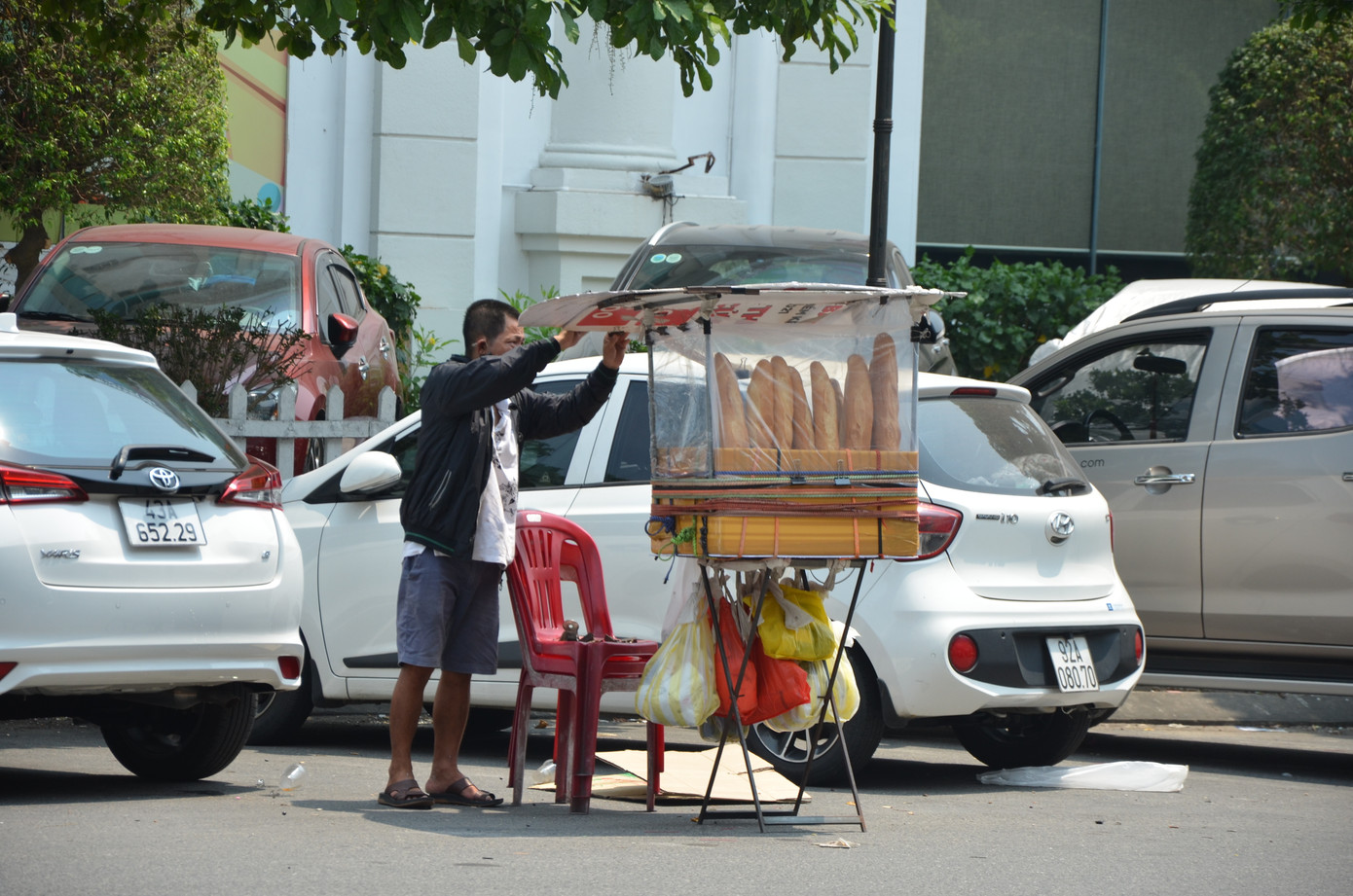 Tại đường Hùng Vương, những người bán bánh mì dạo vốn đã khó khăn nay càng vất vả hơn. Giữa trời nắng gần 40 độ C, họ chỉ có tấm bìa để che nắng.
