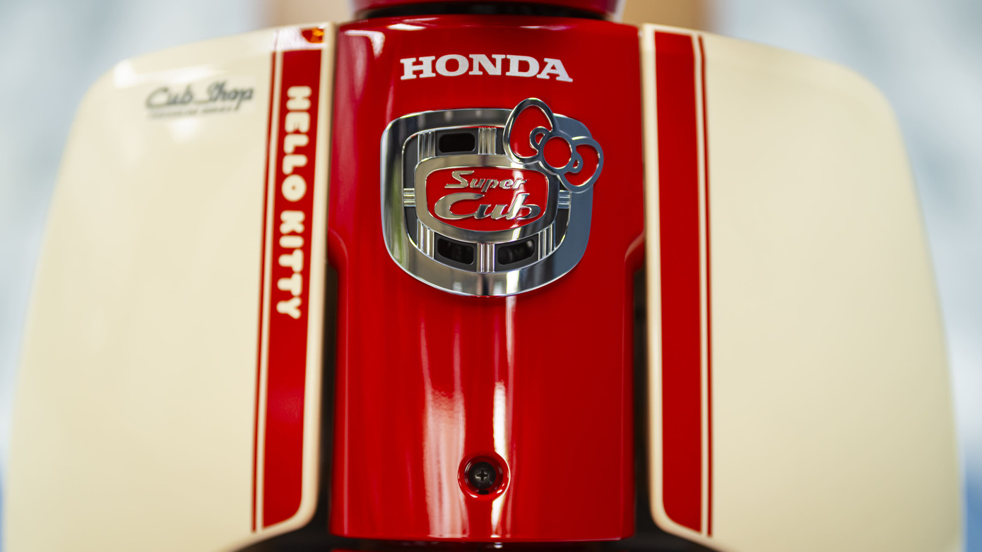 Honda Super Cub Hello Kitty sở hữu phối màu đỏ và be. Logo mèo Hello Kitty xuất hiện trên nhiều vị trí như ốp hông, yếm xe phía trước, bọc yên bằng da, chìa khóa cơ, đồng hồ tốc độ dạng analog kết hợp màn hình kỹ thuật số.