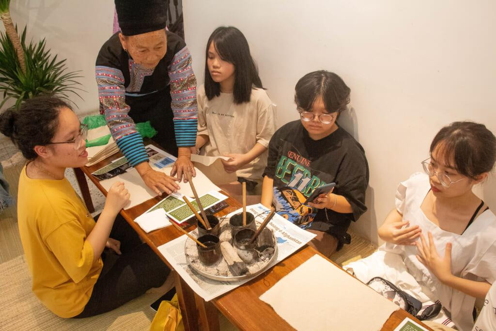 Nghệ nhân dân tộc Mông giàu kinh nghiệm dệt thổ cẩm hướng dẫn các bạn trẻ vẽ sáp ong. Ảnh: Bảo Thu