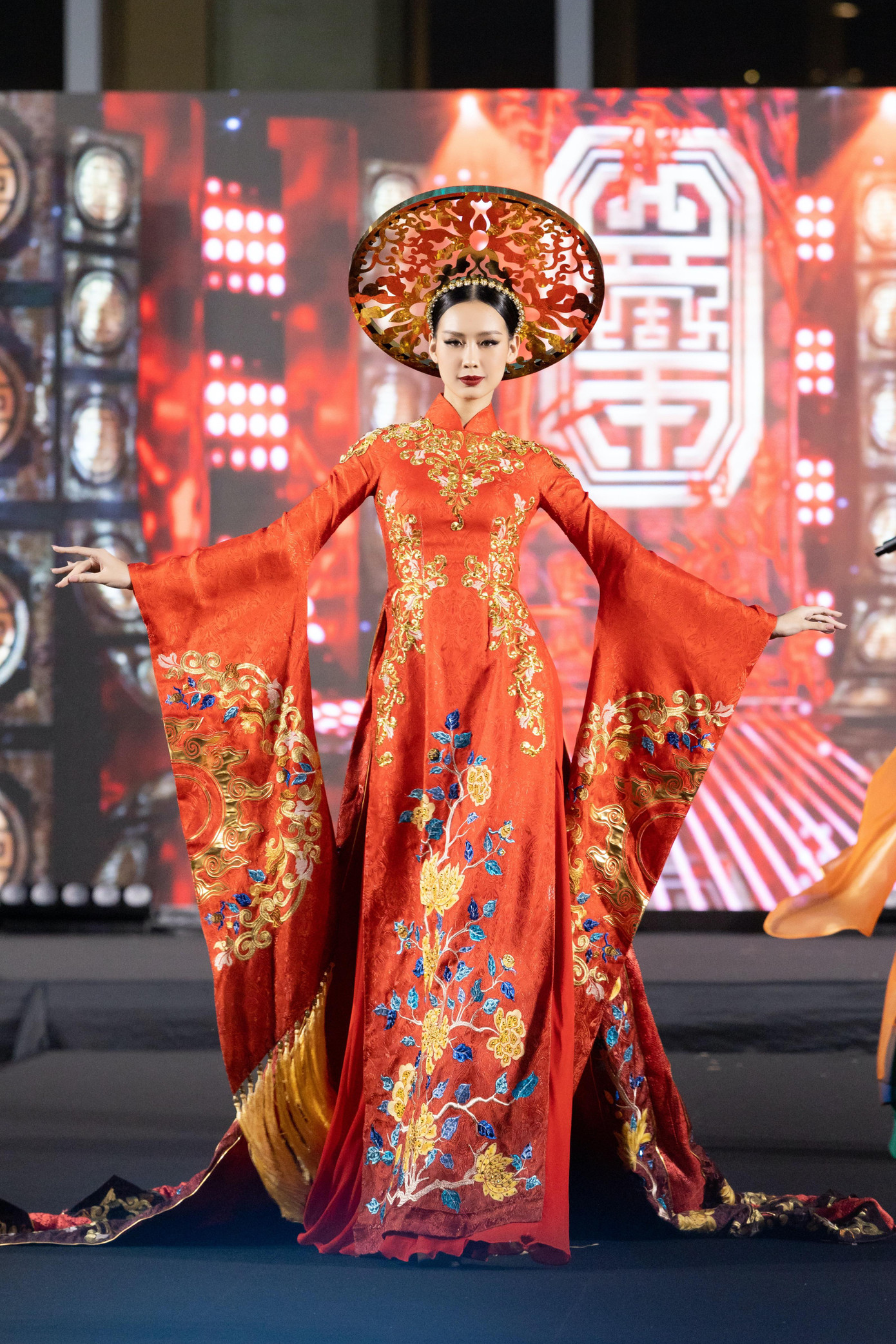 Hoa hậu Bảo Ngọc diện áo dài khăn đóng tông màu đỏ với họa tiết hoa được thêu tỉ mỉ, kỳ công dọc thân áo. Phần tay áo được cách điệu mang lại sự bay bổng và quyền lực cho người đẹp cao 1,85 m khi trình diễn trên sàn catwalk.