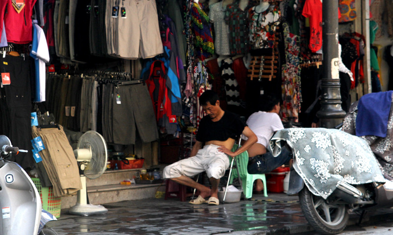 Một chủ hàng quần áo trên phố cổ tránh nóng bằng quạt điện và hắt nước vỉa hè