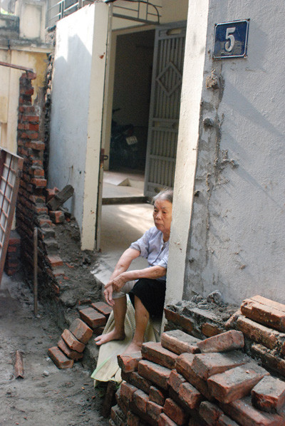 Cụ Lê Thị Dêu ngồi trước cửa ngôi nhà của mình đang bị hàng xóm tiến hành xây bịt lối đi