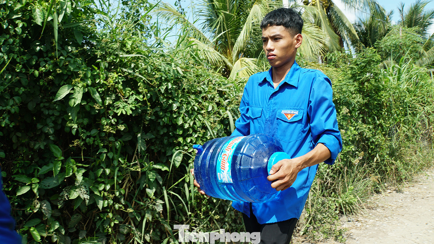 Nhà bà Trinh cách UBND xã hơn cây số, tuy nhiên để đến nơi phải đi trong con hẻm chưa đầy 1m, ngang vừa người, xe gắn máy phải chạy chậm, sâu bên trong. Trong ảnh, đoàn viên thanh niên mang nước đến tận nhà bà Trinh.