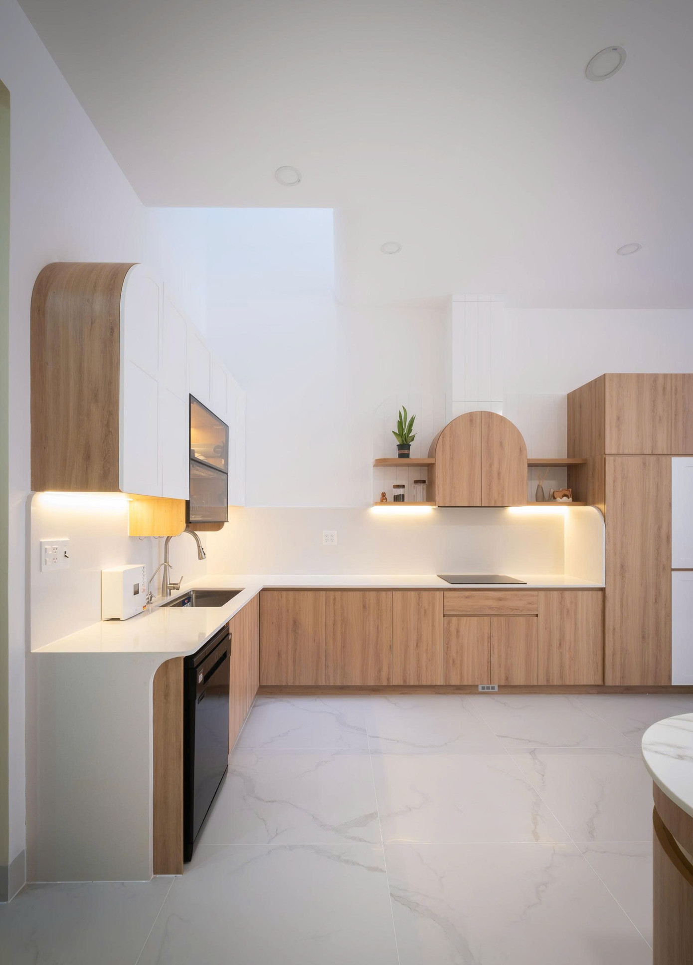 Ngôn ngữ bo tròn tiếp tục được sử dụng triệt để ở khu vực bếp: góc của hệ tủ trên được bo cong mềm mại, máy hút mùi được "giấu" trong một khối trang trí hình vòm cung, tạo sự đồng nhất và duyên dáng.