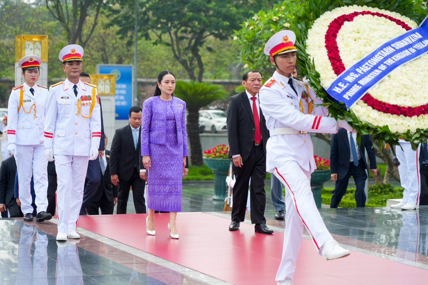 Khi viếng lăng Chủ tịch Hồ Chí Minh và tham gia lễ đón chính thức tại Phủ Chủ tịch, Thủ tướng Thái Lan mặc váy liền thân dáng suông dài và áo khoác ngoài màu tím đậm. Họa tiết trắng trên trang phục thể hiện rõ văn hóa Thái Lan. Ảnh: NBT Connext.