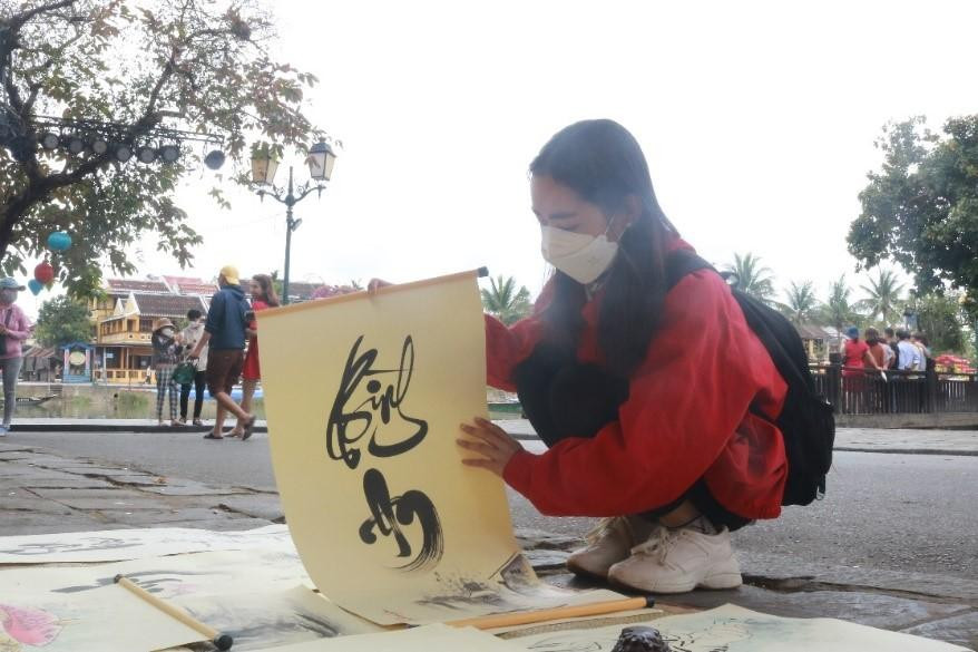 Đồ Vương vui vẻ kể: “Hai năm tôi rồi mới lại thấy cảnh đông đúc như thế này, trong lòng cũng vui hơn nhiều. Vừa cho chữ, vừa ngắm người ta đi chùa Cầu. Từ độ mồng 1 Tết tới nay, hôm nào cũng đông du khách, chủ yếu cho chữ người Việt, còn người Tây họ chỉ ghé xem biểu diễn chữ”. Trong ảnh: Khách du lịch ghé xem và xin chữ - ảnh T.H