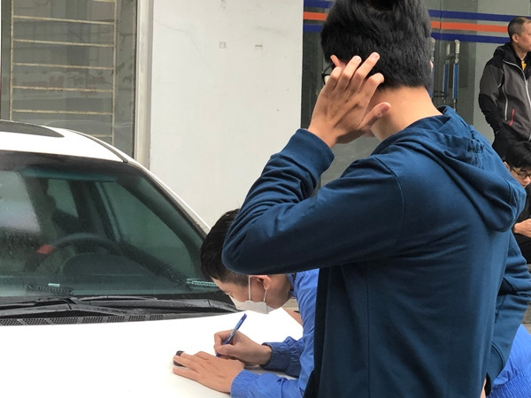 Một phụ huynh (đang viết) lo lắng viết tên tài khoản và mật khẩu của con ra giấy để chạy ra ngoài, đăng nhập rồi lấy giấy báo dự thi và in cho con.