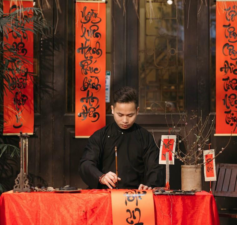 Từ đầu tháng Chạp, Dương Ngọc Tuấn (quê Bắc Giang) đã bận rộn với bút, nghiên, giấy, mực rồi mặc bộ đồ áo dài, khăn xếp đi vẽ những con chữ, câu thơ uốn lượn ở khắp nơi trên địa bàn TP. Hà Nội.