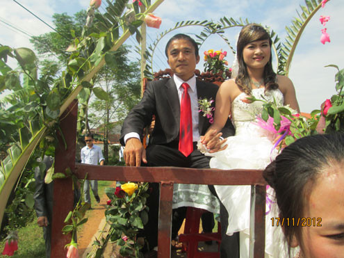 Cô dâu, chú rể nắm tay nhau, hãnh diện ngồi trên xe công nông đi quanh xóm