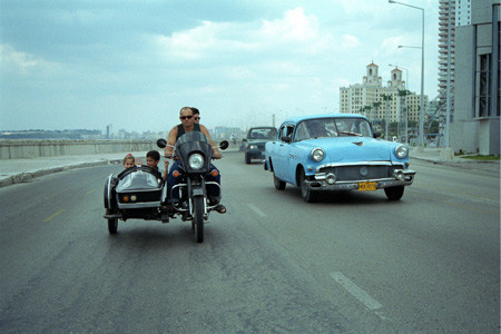 Một người Cu Ba lái xe máy cạnh xe hơi có tuổi đời ít nhất là 50 năm tại đại lộ Malecon ở Havana vào tháng 4 năm 2003