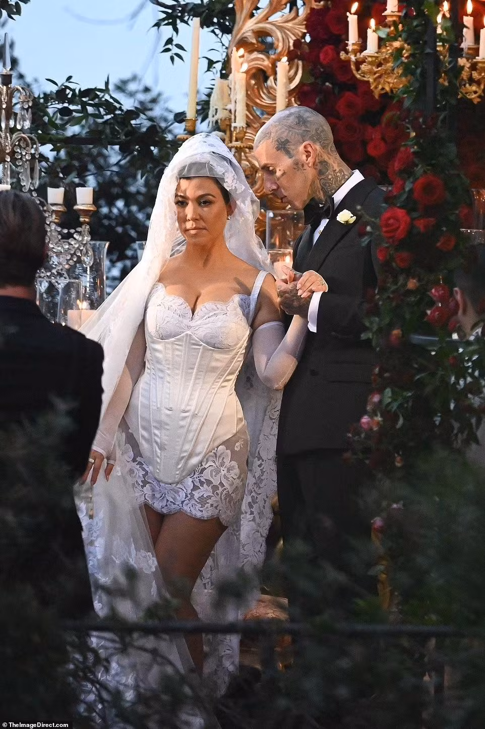 Chị cả nhà Kardashian mặc váy ngắn màu trắng, với chi tiết ren ở phần ngực và chân váy, đội thêm khăn trùm đầu khổng lồ có thêu hoa và hình Đức mẹ Maria. Phụ kiện đi kèm là găng tay trắng và giày đồng bộ lấp lánh. Trong khi đó, Travis diện lễ phục đen thắt nơ cổ điển.