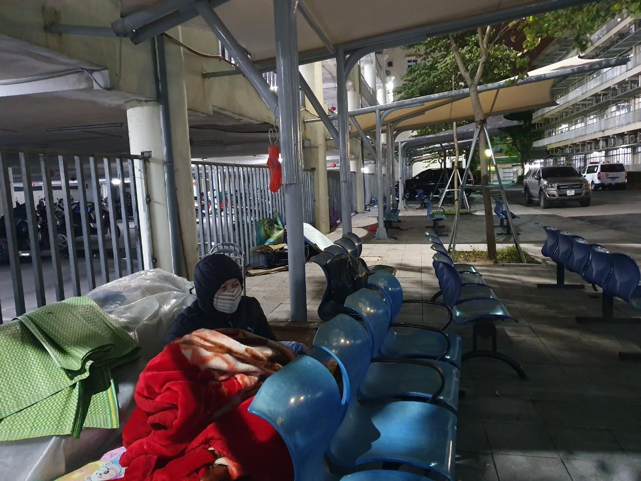 Dưới mái hiên khu nhà xe bệnh viện Bạch Mai, người đàn ông trạc 50 tuổi trùm kín chăn, trở mình liên tục vì gió thốc thẳng vào người. Thấy chúng tôi đến, anh nhổm dậy trò chuyện: “Nằm nhắm mắt để đấy chứ lạnh buốt, lại ngóng tin mẹ, tôi chẳng ngủ được”.