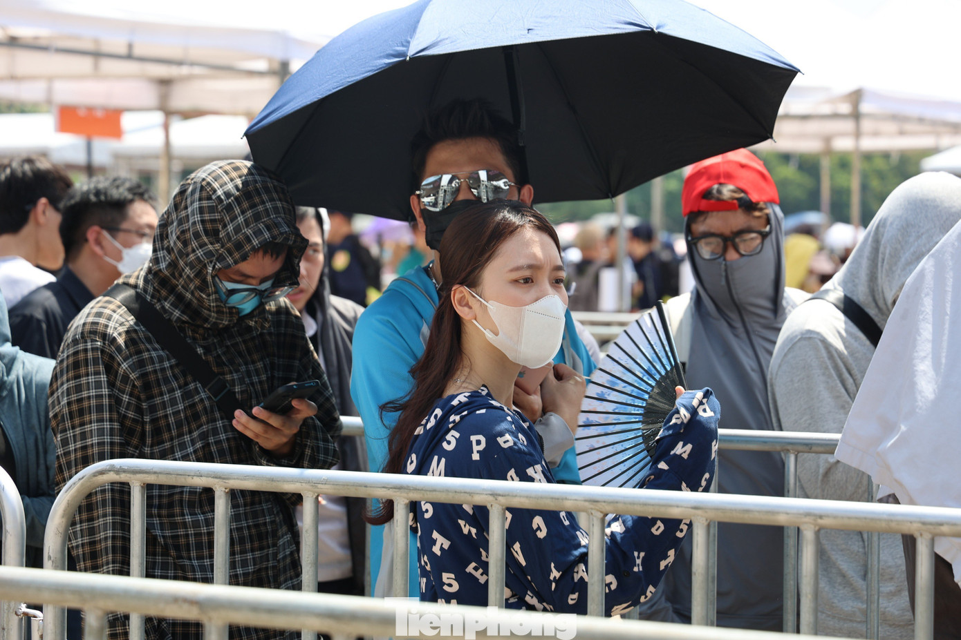 Nhiều người mang ô, mặc kín mít, đeo kính râm đứng xếp hàng dưới cái nắng gay gắt. Ảnh: Như Ý.