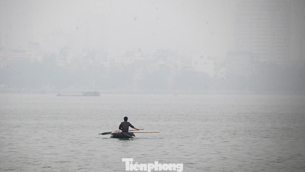 Một người đàn ông chèo thuyền vớt rác trên mặt Hồ Tây mịt mù trong bầu không khí ô nhiễm.