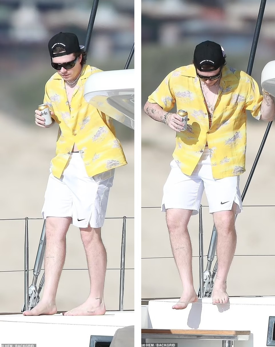 Brooklyn chọn phong cách Hawaii với áo sơ mi cộc tay màu sắc và quần short trắng, có thêm mũ lưỡi trai đội ngược, kính đen và nhiều trang sức khác. Cậu cả nhà Beckham trông trắng "bật tông" khi đứng dưới trời nắng. Ảnh: BackGrid.